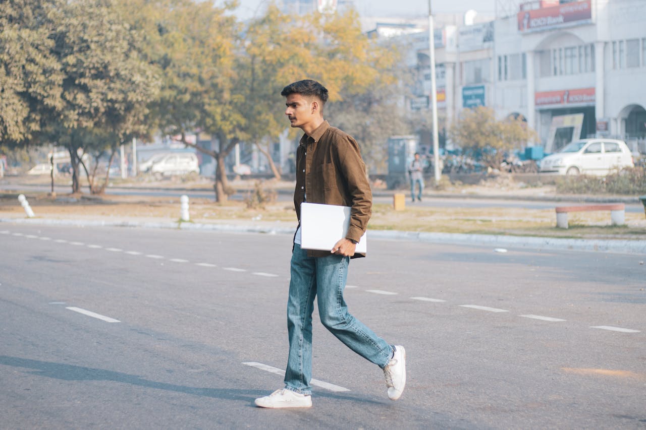 Casual young man crosses a city street holding a laptop, showcasing urban lifestyle.